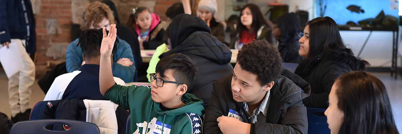 school-aged children at long tables all looking in the same direction. one child has hand raised.